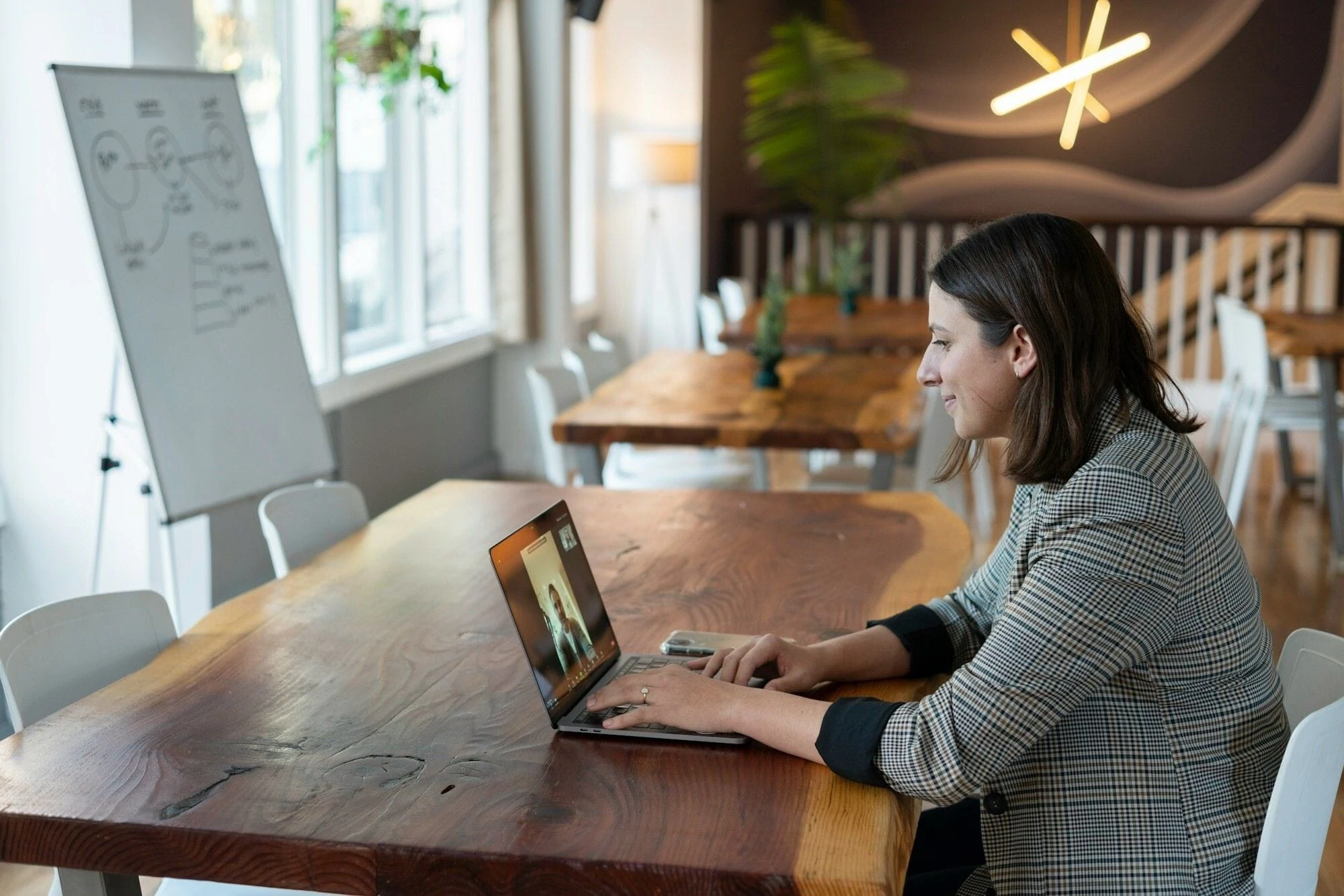 A woman sitting at a table, focused on her laptop, with a cozy workspace in the background.